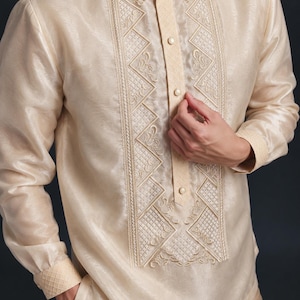 May include: A long-sleeved, champagne-colored barong tagalog shirt featuring a semi-sheer, lustrous fabric. The front panel displays intricate, symmetrical cream-colored embroidery with geometric and floral patterns, a pointed collar, and a button-down placket.
