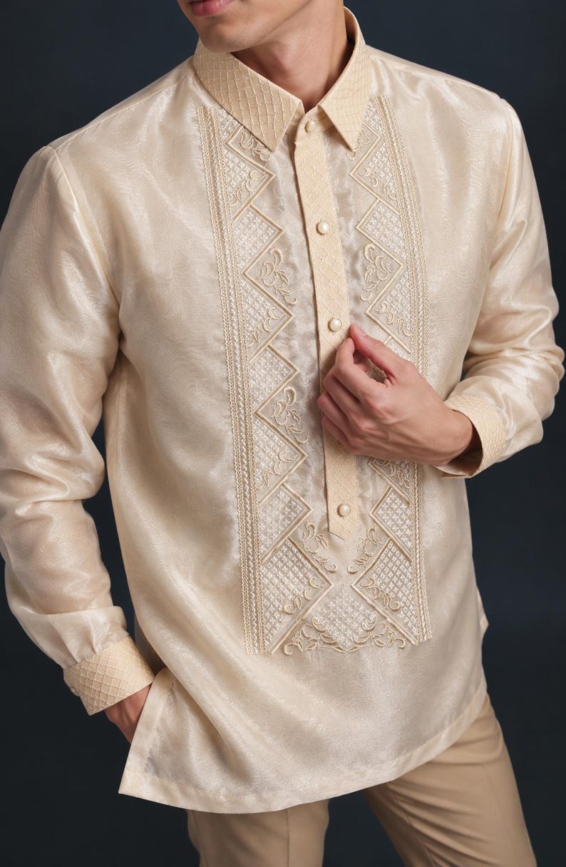May include: A long-sleeved, champagne-colored barong tagalog shirt featuring a semi-sheer, lustrous fabric. The front panel displays intricate, symmetrical cream-colored embroidery with geometric and floral patterns, a pointed collar, and a button-down placket.