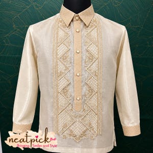 May include: A long-sleeved beige barong tagalog shirt featuring a structured collar, a central button placket, and intricate gold-toned embroidery in geometric and floral patterns along the front panel. The cuffs also feature matching gold embroidery.