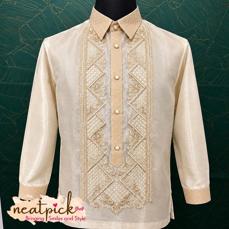 May include: A long-sleeved beige barong tagalog shirt featuring a structured collar, a central button placket, and intricate gold-toned embroidery in geometric and floral patterns along the front panel. The cuffs also feature matching gold embroidery.