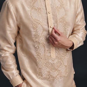 May include: A long-sleeved, champagne-colored barong tagalog shirt featuring a mandarin collar, a button-down placket, and intricate cream-colored embroidery along the front panel, cuffs, and collar with a sheer, textured fabric finish.