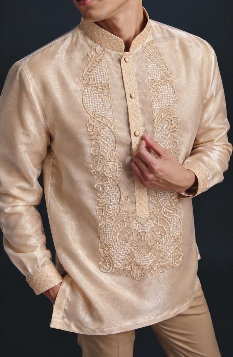 May include: A long-sleeved, champagne-colored barong tagalog shirt featuring a mandarin collar, a button-down placket, and intricate cream-colored embroidery along the front panel, cuffs, and collar with a sheer, textured fabric finish.