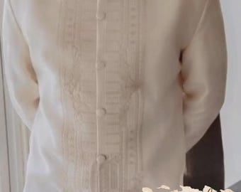 May include: Cream-colored formal shirt with a mandarin collar and button-up front. The shirt features intricate embroidery down the center and is likely made of a lightweight fabric. The shirt is part of a formal wear collection.