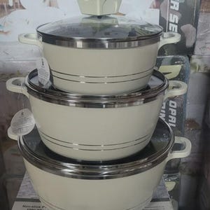 May include: A set of three cream-coloured cooking pots stacked vertically. Each pot features a smooth finish, two side handles, and a silver-toned rim. The top pot includes a clear glass lid with a cream-coloured knob. The pots have horizontal lines.