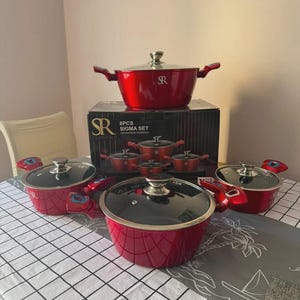May include: An 8-piece SR Essentials Sigma cookware set featuring metallic red pots with silver-toned handles and clear glass lids. The set includes various sizes of pots, each with a dark speckled interior, displayed with their original black box.