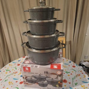 Peut inclure: Une pile de quatre casseroles en céramique gris foncé moucheté avec des bords et des poignées argentés, surmontée d'un couvercle en verre. Les casseroles reposent sur une boîte en carton marquée Delight Leo Ceramic Set avec le texte 8pcs.