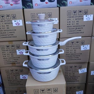 Op de afbeelding: Gestapelde witte kookgerei set met getextureerde buitenkant en zilveren randen, met het label "IBRAA". De afbeelding toont meerdere sets, met dozen met "COOKWARE SET" en productdetails. De dozen zijn gestapeld.