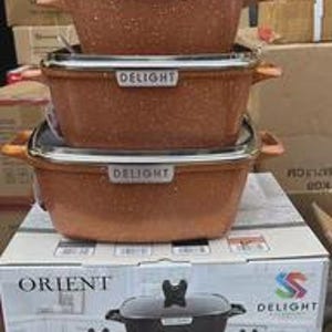 May include: A set of three copper-colored non-stick casserole dishes with lids, stacked on top of a box. Each dish has the word "DELIGHT" on the front. The box below displays the "ORIENT" and "DELIGHT" logos and the words "DIE-CAST CASSEROLE SET".