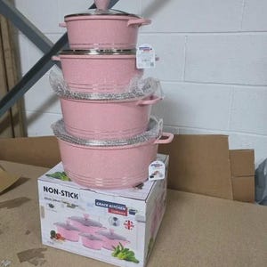 Op de afbeelding: Een stapel roze non-stick kookgerei met zilveren randen en handvatten. De set bevat potten en deksels, sommige verpakt in beschermend plastic. De doos hieronder toont de merknaam "Grace Kitchen Cookware" en het woord "Non-Stick".