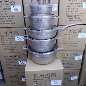 Könnte beinhalten: Ein Stapel neuer, champagnerfarbener Kochgeschirrsets mit strukturierten Oberflächen, Deckeln und Griffen. Die Sets sind auf Kartons gestapelt. Die Kartons tragen Produktinformationen. Maße: 36x36x32,5 cm.