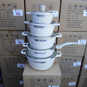 Könnte beinhalten: Ein Stapel cremefarbener Kochtöpfe mit Deckeln und Griffen, beschriftet mit "IBRAA", ist zu sehen. Die Töpfe haben ein strukturiertes Korbgeflecht-Design. Kartons mit Produktinformationen, einschließlich Abmessungen von 36x36x32,5 cm, sind ebenfalls sichtbar.