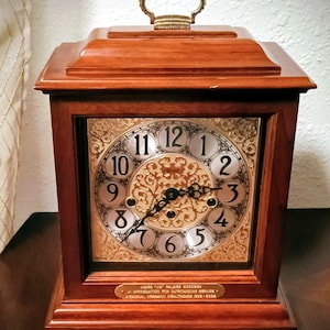 May include: A wooden mantel clock with a square face and a gold-colored clock face. The clock has a handle on top and a small plaque at the bottom with text. The clock face has black hands and silver numerals.