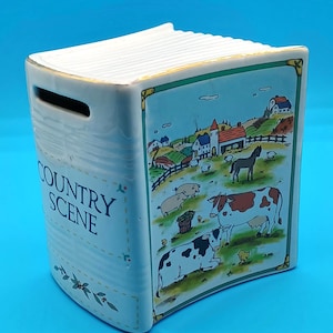 May include: A ceramic bank shaped like a book, featuring a "Country Scene" design. The illustration depicts a rural landscape with cows, pigs, a horse, and a farmhouse. The bank has a slot for inserting money.