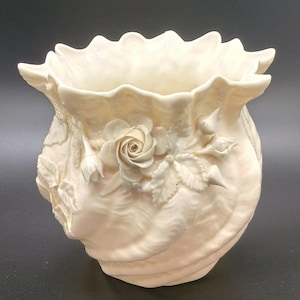 May include: A cream-colored ceramic vase with a ruffled rim and a textured, shell-like body. The vase features a raised floral design with a central rose and several buds and leaves. The overall design is elegant and decorative.