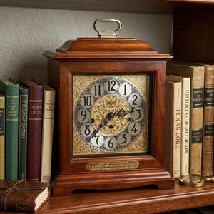May include: A wooden mantel clock with a square face, black Arabic numerals, and ornate gold-toned metal detailing. The clock features a top handle and a brass plaque at the base with the text: JAMES "ZH" PALMER WOODSON IN APPRECIATION FOR OUTSTANDING SERVICE MEMORIAL HERMANN HEALTHCARE 1906-0000.