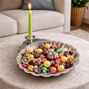 May include: A lit green candle in a silver candlestick holder sits next to a decorative silver serving dish filled with assorted chocolates. The chocolates are wrapped in gold, red, silver, and green foil.