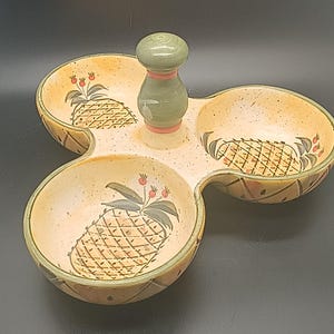 May include: A ceramic serving dish with three sections and a central handle. The dish is beige with a speckled finish and features hand-painted pineapple designs. The handle is green with a pink accent. The dish has a decorative border.