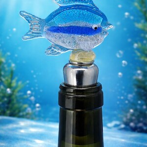 May include: A decorative wine bottle stopper featuring a blue glass fish design. The fish has a blue stripe and is mounted on a silver and black stopper, atop a green glass bottle. The background is a blue underwater scene.