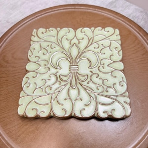 May include: A square ceramic tile featuring a pale green glaze with a raised, dark-toned fleur-de-lis motif centered amidst intricate, symmetrical scrollwork patterns that extend to the scalloped edges of the piece.