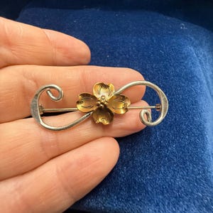 May include: A silver-toned metal brooch featuring a central four-petaled flower with a warm bronze finish. The flower is flanked by two symmetrical, curved silver-toned metal scrolls that extend outward to form an elongated, horizontal shape.
