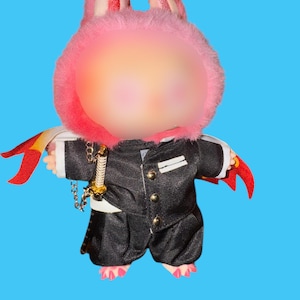 May include: A plush doll wearing a black suit with gold buttons, a white shirt, and black pants. The doll has a pink bunny-eared hood and a small sword. Red and yellow accents are visible on the doll's arms. The background is a solid blue.