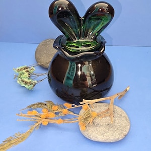 May include: A dark green and black glass vase with a distinctive, heart-shaped top. The vase has a rounded base and is set on a blue surface, with decorative stones and dried flowers nearby.