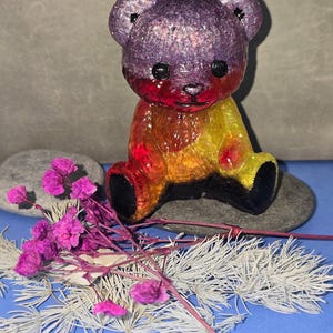 May include: A colorful, glittery teddy bear figurine with a gradient of purple, red, yellow, and black. The bear is seated on a gray stone, with pink flowers and white foliage in the foreground. The background is a solid blue surface.