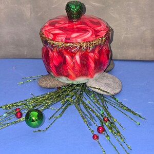 May include: A red and white jar with a lid, featuring gold trim and a green glitter knob. The jar is surrounded by green glittery sprigs, red berries, and a green bauble. The jar is approximately 10 cm in height.