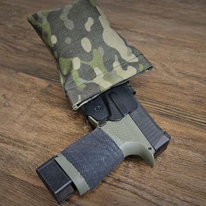 Puede incluir: Una pistola semiautomática con corredera negra y armazón verde oliva está parcialmente cubierta por una funda de tela de camuflaje. La empuñadura está envuelta en un material negro texturizado.