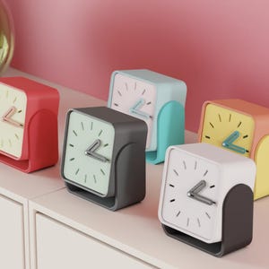 May include: A collection of five colorful, square-faced analog clocks. Each clock has a different color scheme: red, gray, light blue, yellow, and white. The clocks are displayed on a white surface against a pink background.