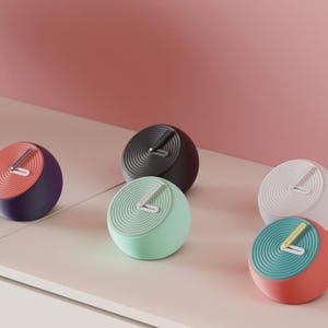 May include: A selection of modern, spherical clocks in colours such as red, teal, black, white, and mint green. Each clock has a contrasting coloured face and a single hand. The clocks are displayed on a white surface against a pink background.