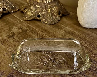 May include: Clear glass butter dish with a floral design on a wooden surface. The dish has a rectangular shape with a lid and handles. Decorative items are in the background.