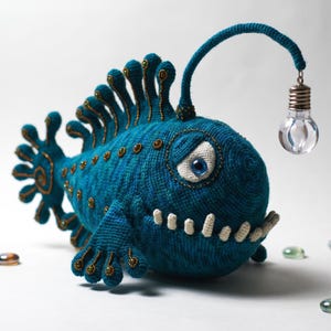 Puede incluir: Un pez rape de ganchillo en color azul cerceta con una aleta dorsal prominente, un ojo grande blanco con pupila azul y una hilera de dientes blancos pequeños. Un tallo largo y fino de ganchillo sale de su cabeza y termina en una bombilla.