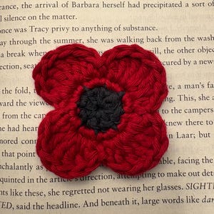 May include: A handmade, crocheted red poppy flower with a black center, resting on a book page. The four-petal flower is a symbol of remembrance. The text on the page is partially visible.