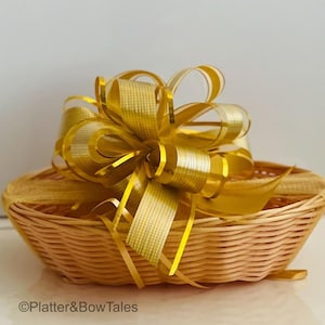 May include: A small, woven wicker basket with a large, decorative gold bow. The bow is made of multiple layers of gold and sheer ribbon. The basket is empty and sits on a white surface. The text "©Platter&BowTales" is visible.