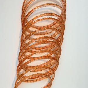 May include: A collection of transparent orange bangles, adorned with tiny gold-coloured beads. The bangles are stacked in a spiral formation, highlighting their intricate design and warm hue. The beads provide a subtle shimmer to the bracelets.