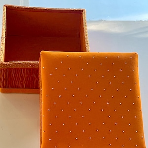 May include: Open orange square box with a matching lid. The lid is covered in small, evenly spaced, silver-colored embellishments. The box's interior is orange, and the rim is decorated with gold trim and a red patterned border.
