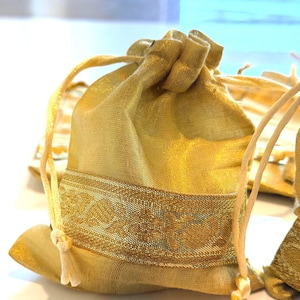 May include: Gold drawstring pouch with a decorative band. The pouch is made of a shimmering gold fabric and features a decorative band with an intricate pattern. The drawstring is a light beige color. The pouch is likely used for small gifts or jewelry.