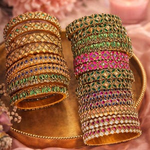 May include: Stacks of gold bangles with intricate designs and colorful gemstones. The bangles feature green, pink, blue, and white stones, arranged in floral and geometric patterns. The jewelry is displayed on a gold tray.