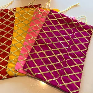 May include: Four colorful drawstring bags in red, yellow, pink, and magenta. Each bag features a gold diamond grid pattern. The bags are made of a soft, velvety material and have gold drawstrings.
