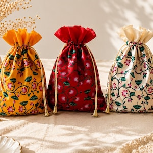 May include: Three drawstring pouches in yellow, red, and cream, each featuring a repeating floral pattern of pink lotus flowers and green leaves. The bags are cinched at the top with thin, light-coloured cords that have small metallic bell-shaped ends.