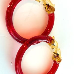 May include: Two circular red glass bangles featuring clear amber-colored glass elephant head motifs at the ends. The bracelets have a smooth, translucent finish and are designed with the elephant trunks meeting to form the closure of the hoop.