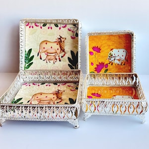 May include: Four decorative metal trays with ornate silver frames. The trays feature colorful cow illustrations on a yellow and floral background. The trays are square and rectangular, with a matching design.