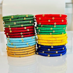 May include: Stacks of colorful bangles in green, red, blue, yellow, and gold. Each bangle is adorned with small, gold-colored floral accents. The bangles are stacked neatly, showcasing a variety of vibrant colors and textures.