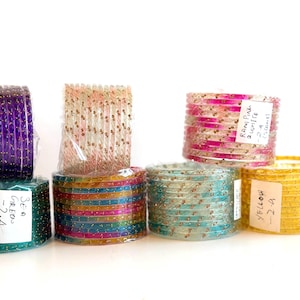 May include: A selection of colourful glass bangles, including purple, green, pink, and yellow. Each stack is wrapped in clear plastic and decorated with small, gold-coloured embellishments. Some stacks have labels with handwritten text.