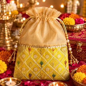 May include: A yellow drawstring pouch featuring a diamond-patterned embroidered design with small colourful floral motifs and gold metallic thread. The top section is made of sheer gold fabric with matching gold cords finished with pearl-like beads.