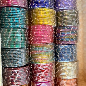 May include: A selection of vibrant glass bangles in shades of red, blue, green, and gold. Each stack of bangles is wrapped in clear plastic. The bangles are decorated with small, decorative embellishments.
