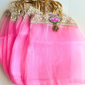 May include: A collection of bright pink organza gift bags featuring gold metallic handles. Each bag is adorned with a wide, decorative silver and gold embroidered trim along the top edge, accented by a small pink lotus flower charm with gold beads.