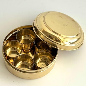 May include: A round, golden brass spice box with a removable lid. The box is divided into seven compartments, each designed to hold different spices. The lid is slightly ajar, revealing the interior compartments. The box is on a white surface.