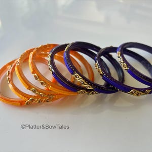May include: A collection of colorful bangles. The set includes orange and purple bangles with gold-colored embellishments. The bangles are arranged in a row, showcasing their intricate designs and vibrant hues.